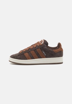 Zapatilla de ante marrón con tres rayas de acento marrón, puntera redonda, cordones marrones y suela blanca. Detalle del logo en la lengüeta y el talón.