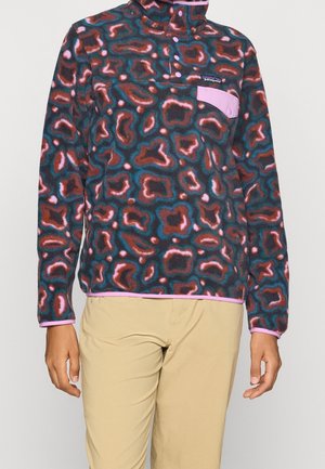 Bluza polarowa z wielokolorowym abstrakcyjnym wzorem, kieszenią na piersi na zatrzask z różową klapką oraz różowym obszyciem, noszona z beżowymi spodniami.