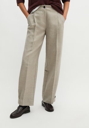 Personne portant un pantalon beige à jambes larges avec des revers, des mocassins marron foncé et un pull bordeaux, les mains dans les poches, debout sur un fond clair.