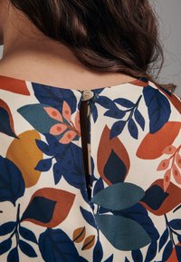 Blouse à motif floral avec fermeture au dos en forme de goutte et bouton, en couleurs crème, bleue, orange et verte. Texture de tissu douce.