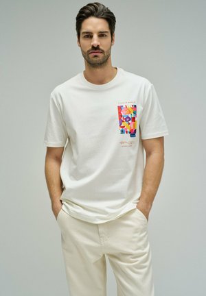 Hombre con camiseta blanca con diseño abstracto colorido en el pecho izquierdo, de pie con las manos en los bolsillos contra un fondo liso.