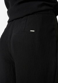 Black pants with a textured fabric, featuring a front pocket and a small silver brand tag. The waistband is elasticized for comfort.