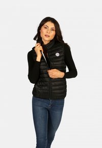 Gilet rembourré noir avec un col haut, fermeture éclair sur le devant et un logo circulaire sur la poitrine gauche. Poches latérales profondes et texture légère.