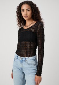 Svart langermet topp laget av teksturert gjennomsiktig stoff med bølgemønstre, kombinert med lys blå jeans med klassisk knappelukking.