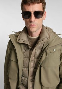 Giacca da esterno verde oliva con molte tasche, indossata sopra un gilet imbottito beige. Il modello indossa occhiali da sole scuri, evidenziando un design elegante.