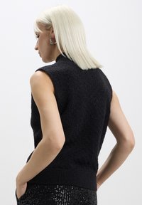 Top senza maniche nero in maglia con finitura testurizzata. Collo rotondo, orlo a costine e silhouette aderente. Modello che indossa pantaloni con paillettes.