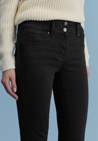 Schwarze High-Waist-Jeans mit Slim-Fit, ausgestattet mit zwei vorderen Taschen und zwei Knopfverschlüssen. Weicher Baumwoll-Mischstoff mit glatter Textur.