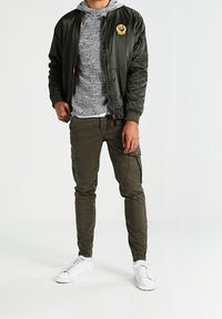 Groene bomberjack met badge, grijze hoodie, olijfgroene cargo broek en witte sneakers. Gladstoffen textuur en getailleerd ontwerp.