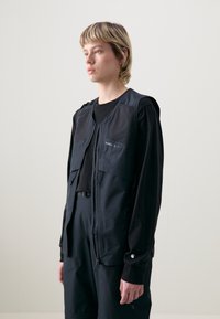 Crna zip-up prsluk s dva prednja džepa, lagana, teksturirana tkanina i elastični detalji. Nositi preko crne košulje s dugim rukavima.