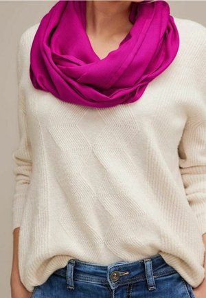 Kvinde iført en cremet hvid tekstureret trøje, knaldmagenta infinity-sjal og blå jeans med hænderne i lommerne.