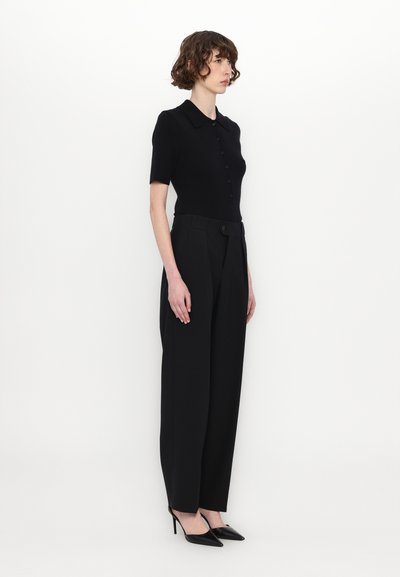Haut noir à manches courtes et col avec détail de boutons, associé à un pantalon noir taille haute à larges jambes et des escarpins noirs pointus.
