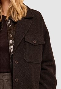 Abrigo de mezcla de lana oscura con un acabado texturizado, que presenta un gran bolsillo, solapa y botones marrones para el cierre. La capa inferior muestra un material brillante.