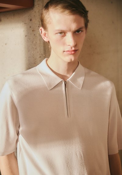 Lichtroze polo met korte mouwen en een ritskraag, gemaakt van zachte, gestructureerde stof. Het ontwerp heeft een schone, minimalistische esthetiek.