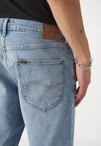 Gaiši zili džinsi ir aprīkoti ar brūna ādas uzlīmi ar uzrakstu "Lee" aizmugures jostas vietā, un sānu skats uz kabatu ar redzamām šuvēm.