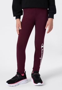 Burgundowe legginsy z elastycznej tkaniny, z białym logo "Champion" wzdłuż lewej strony, w połączeniu z czarnymi butami sportowymi z różowymi akcentami.