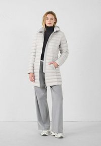 Lichtgrijze pufferjack met rits aan de voorkant, grote capuchon en verticale stiksels; gedragen over een zwarte coltrui en grijze wijde broek.