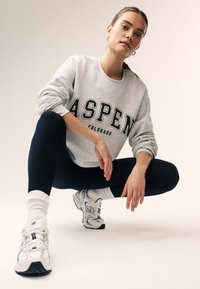 Giovane donna accovacciata che indossa una felpa grigia "Aspen Colorado", leggings neri, calzini bianchi e sneakers bianche New Balance su uno sfondo semplice.