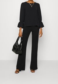 Pantalons noirs évasés avec des rayures verticales, associés à un haut noir à manches longues avec un ourlet volant. Accessoirisé avec un sac à main noir texturé.