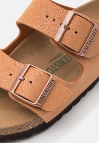 Birkenstock sandaal met een tan gekleurde textuur bovenkant, twee verstelbare banden, metalen gespen en een bruine kurken binnenzool.