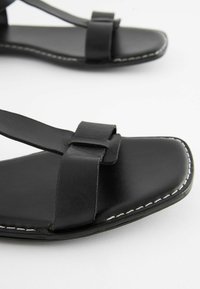 Mustat nahka-sandaalit, joissa on litteät pohjat ja minimalistinen muotoilu. Ominaisuuksiin kuuluu kaksi hihnaa, jotka on yhdistetty nauhalla, ja valkoiset ompelut.