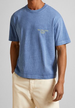 Camiseta de algodón azul con mangas cortas y cuello redondeado. Presenta texto bordado en amarillo en el pecho. Combina con pantalones beige.