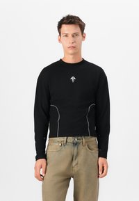 Sort langærmet skjorte med et struktureret mønster, der har et lille hvidt logo og kontrasterende hvid syning langs siderne, parret med slidte jeans.