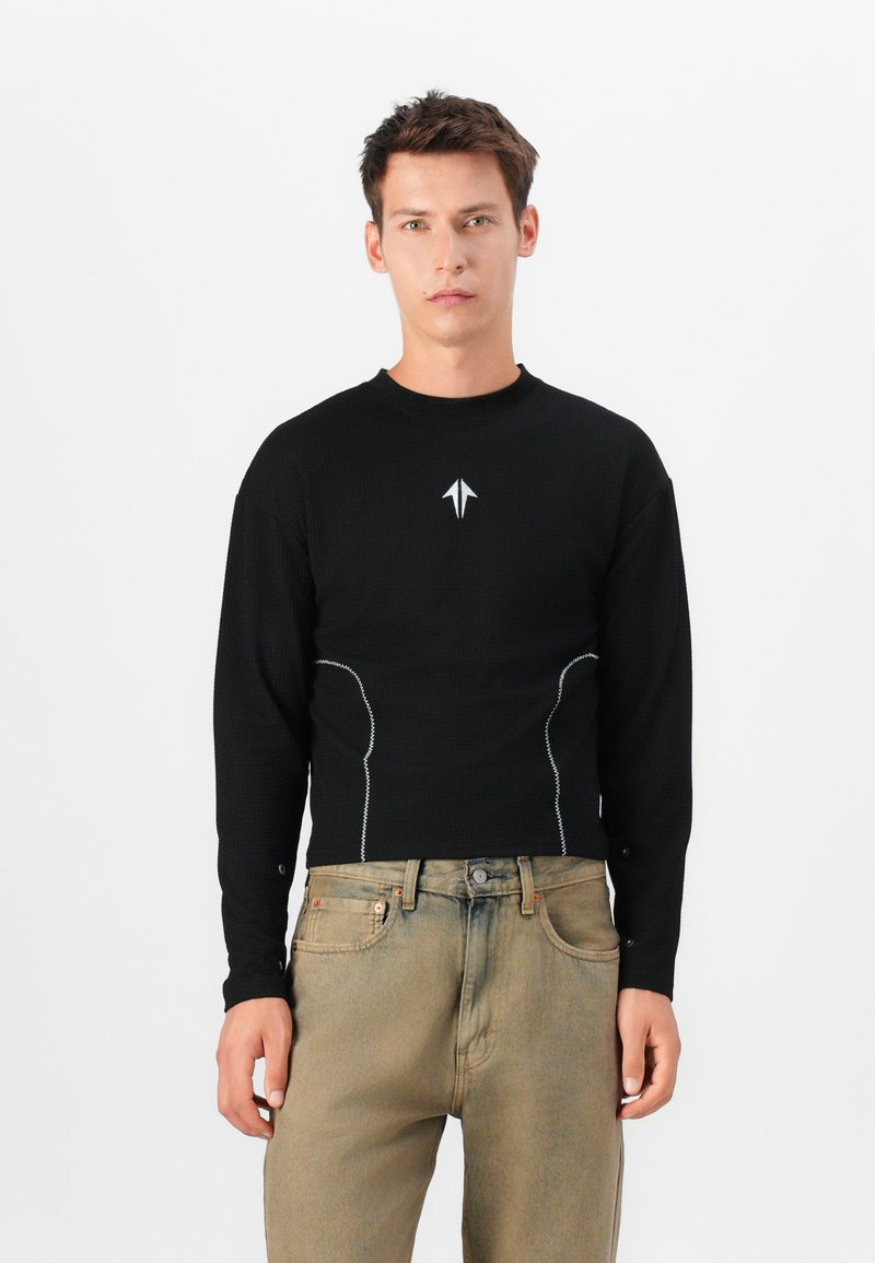 Sort langærmet skjorte med et struktureret mønster, der har et lille hvidt logo og kontrasterende hvid syning langs siderne, parret med slidte jeans.