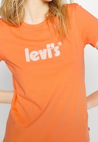 Camiseta de algodón naranja con cuello redondo y mangas cortas. Presenta un logo blanco de "Levi's" impreso en el frente. Textura suave.