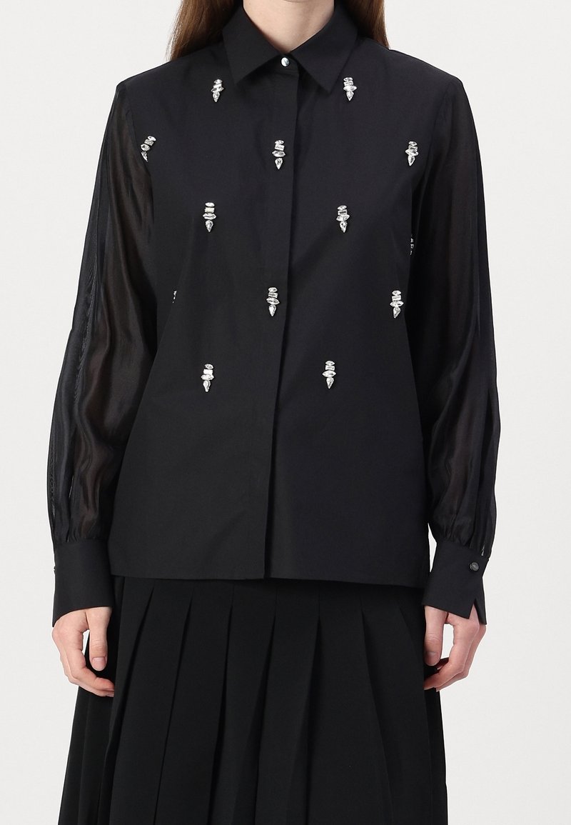 Blouse noire à manches longues avec manches transparentes et ornements en perles blanches dispersées, associée à une jupe plissée noire.