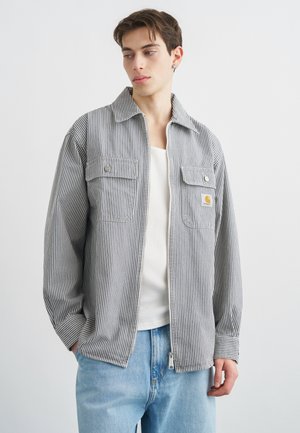 Jeune homme portant une veste zippée rayée grise et blanche par-dessus un t-shirt blanc et un jean bleu clair, regardant sur le côté.