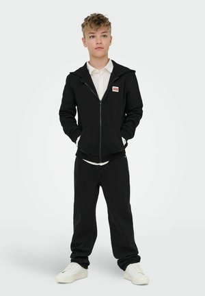 Niño de pie con las manos en los bolsillos, vistiendo una sudadera negra con cremallera, camisa blanca con cuello, pantalones negros y zapatillas blancas sobre un fondo liso.