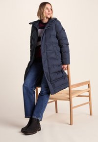 Woman wearing a navy long puffer coat, striped sweater, blue jeans, and black boots sitting on the edge of a wooden chair with woven seat.