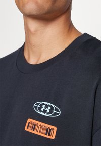 Námořnická modrá bavlněná tričko s světle modrým logem a oranžovým textovým grafickým prvkem 'UNDER ARMOUR' na přední straně. Crew výstřih, krátké rukávy.
