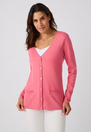 Frau trägt eine rosa Strickjacke mit Knöpfen und zwei vorderen Taschen sowie weiße Hose, steht vor einem schlichten hellen Hintergrund.