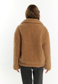 Brun, fuzzy, tekstureret jakke med en bred krave og sænkede skuldre, parret med mørkegrå bukser. Ingen synlige lukninger eller mønstre.