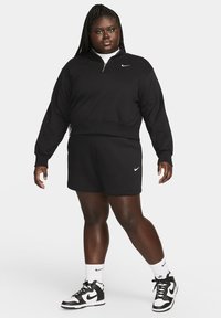 Fekete Nike pulóver negyedcipzárral, fekete rövidnadrággal párosítva. Figyelemre méltó fehér Nike logó mindkettőn. Fekete-fehér sportcipő.