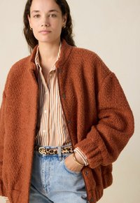 Veste en polaire orange rouille avec une finition texturée, équipée de boutons pression. Portée sur une chemise à rayures à col, assortie à un jean en denim clair.