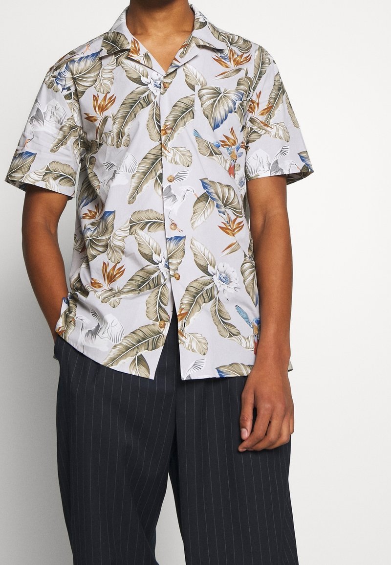 Camicia a maniche corte con sfondo bianco e un motivo di foglie e fiori tropicali in tonalità di verde, arancione e blu; chiusura con bottoni.