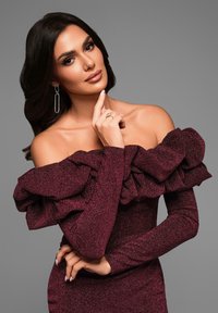 Burgundy off-shoulder klänning med volanger, långa ärmar och en texturerad tyg. Utrustad med silvriga hängande örhängen och framträdande ringar.