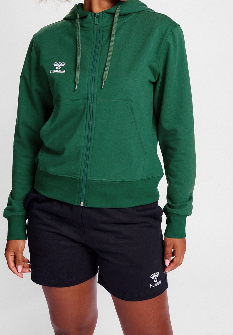 Sudadera verde con cremallera, hecha de material suave, con un bolsillo tipo canguro, cordones a juego y un logotipo en el pecho. Combinada con pantalones cortos negros.