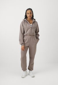 Gecombineerde hoodie en joggers in lichtbruin fabric, voorzien van een rits, een elastische tailleband en een ontwerp met omgeslagen enkels.