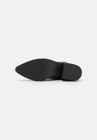 Black leather shoe with a pointed toe, flat sole, and strap detail. Textured rubber outsole features horizontal ridges for grip.