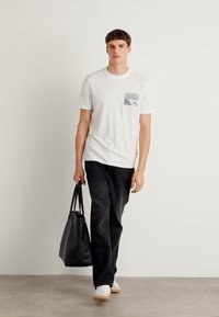Hvid t-shirt med korte ærmer og en grå lomme, sorte bukser og hvide sneakers; holder en sort tote bag; ensfarvet baggrund.