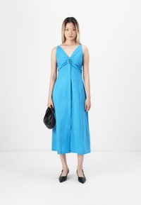 Kvinne stående i en blå ermeløs midikjole, holder en svart handveske, iført svarte spisse slingback-heler mot en hvit bakgrunn.