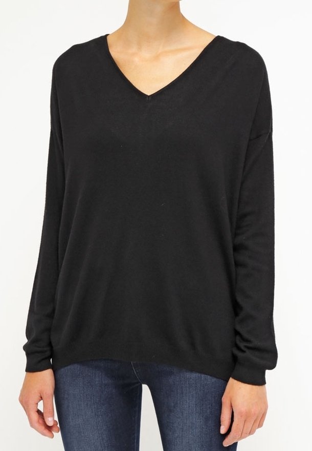 Femme portant un pull ample noir à col en V et manches longues, associé à un jean bleu foncé, debout devant un fond blanc uni.