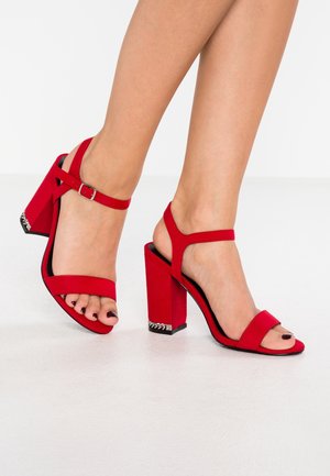 Punaiset korkokengät, joissa on leveä nauha varpaiden yli ja nilkkahihna soljella, tukevalla korolla. Teksturoitu pinta.