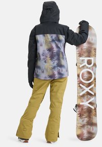 Giacca da snowboard nera con motivo viola a alberi, abbinata a pantaloni gialli. Grande scritta bianca "ROXY" sulla tavola da snowboard.