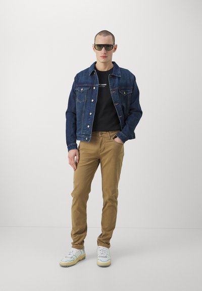 Jachetă din denim, tricou negru, pantaloni bej și adidași albi cu accente galbene. Model purtând ochelari de soare negri, stând în fața unui fundal simplu.