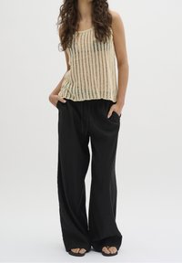 Nainen, joka on pukeutunut beigestä olkaimettomaan neulepuseroon, löysiin mustiin housuihin, joissa on taskut, ja mustiin flip-flop-sandaaleihin, seisoo kädet taskuissa.