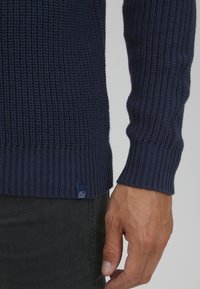 Navy pletena pulover z teksturiranim vzorcem, rebrastimi manšeti in robom ter majhnim modrim oznako na spodnji levi strani.